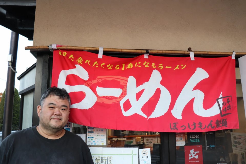 ぼっけもん製麺の店主と、店頭の「らーめん」の赤いのれん